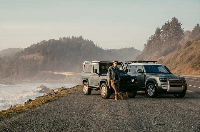 John Mayer poses with a vintage Defender and the New Defender on a California shoreline.