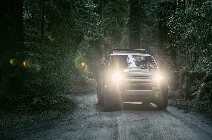 The New Defender driving with headlights on in a dark forest.
